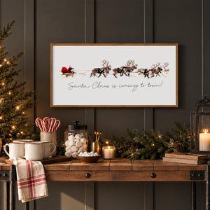 Puede incluir: Un letrero de pared rectangular con marco de madera que presenta un gr&aacute;fico de Santa Claus en un trineo tirado por renos y el texto Santa Claus is coming to town! El letrero se muestra en una pared oscura sobre una mesa de madera con decoraci&oacute;n.