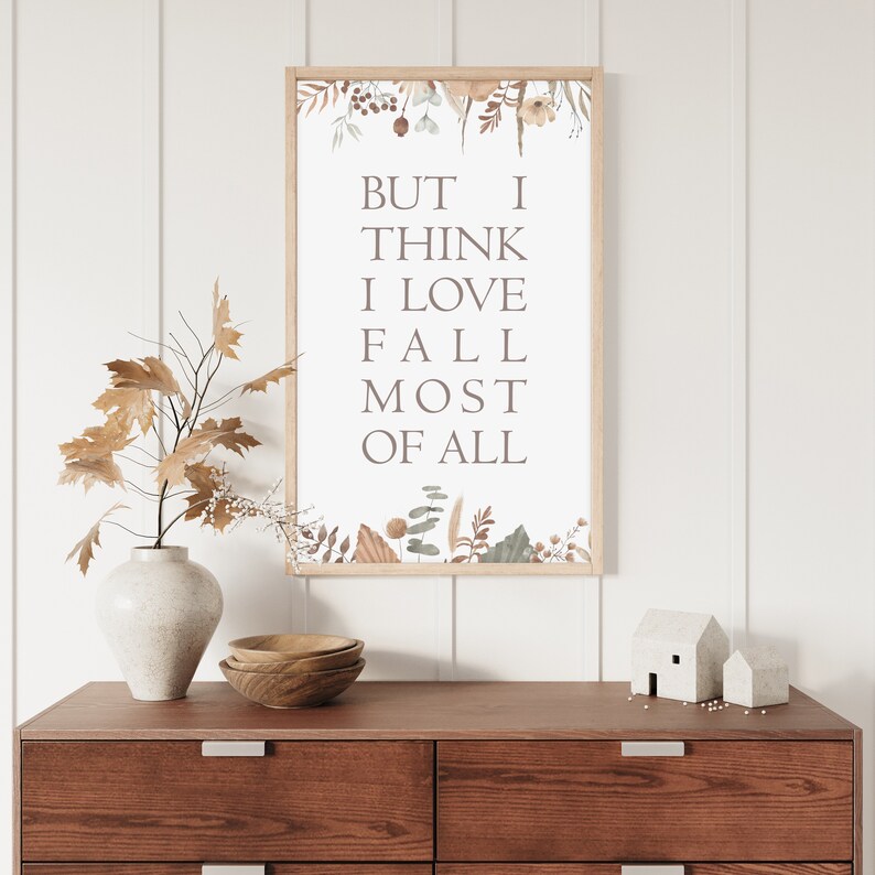Puede incluir: Un cuadro rectangular con marco de madera clara que contiene el texto BUT I THINK I LOVE FALL MOST OF ALL en una fuente serif. El dise&ntilde;o est&aacute; decorado con ilustraciones estilo acuarela de hojas secas, bayas y follaje en la parte superior e inferior.