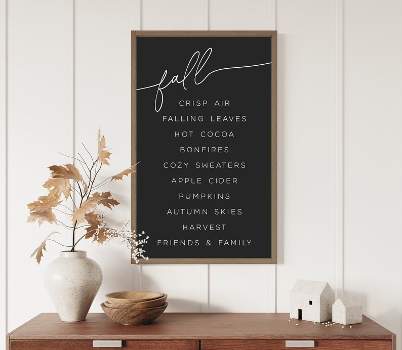 Op de afbeelding: Een zwart geframed schoolbord met witte tekst die "fall" in een cursief schrifttype leest. Het bord vermeldt de volgende woorden: frisse lucht, vallende bladeren, warme chocolademelk, kampvuren, gezellige truien, appelcider, pompoenen, herfstlucht, oogst, vrienden en familie.
