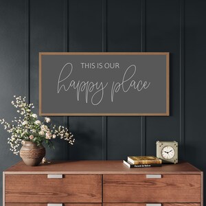 May include: A rectangular wall sign with a light brown wooden frame and a dark gray background. The text "THIS IS OUR" is printed in a simple sans-serif font, while the word "happy place" is written below in a white, flowing cursive script.