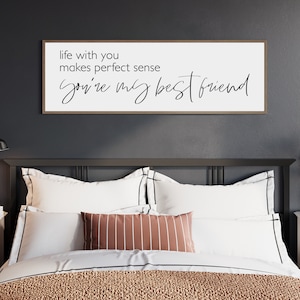 May include: A white sign with a black wood frame hanging above a bed with a brown and white patterned comforter. The sign reads "life with you makes perfect sense you're my best friend".