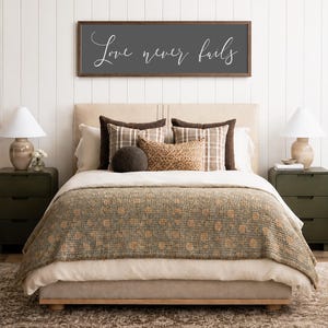 May include: A bedroom scene featuring a bed with a neutral-toned headboard and bedding. A decorative sign above the bed reads "Love never fails" in white script. The bed is adorned with patterned pillows and a textured throw blanket. Two green nightstands flank the bed, each with a lamp.