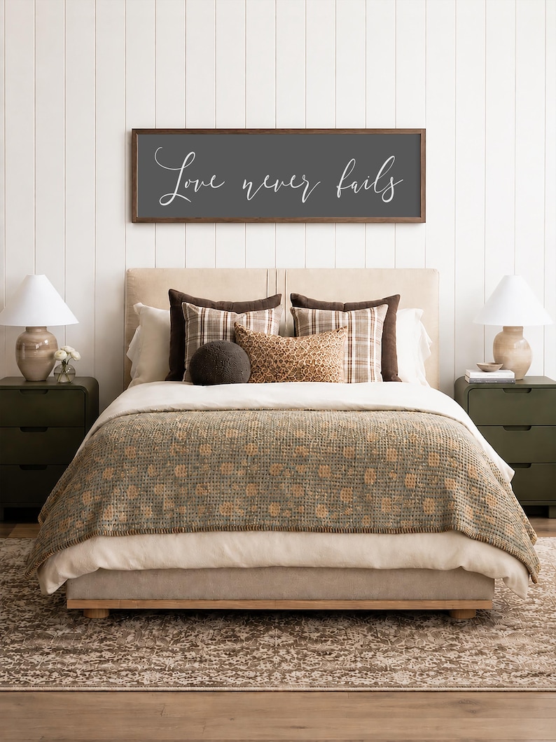 May include: A bedroom scene featuring a bed with a neutral-toned headboard and bedding. A decorative sign above the bed reads "Love never fails" in white script. The bed is adorned with patterned pillows and a textured throw blanket. Two green nightstands flank the bed, each with a lamp.