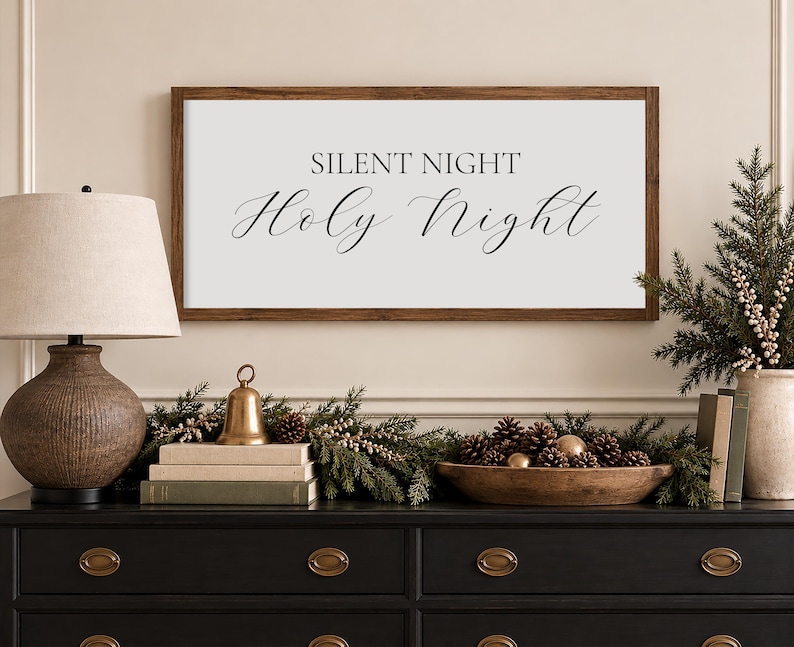 May include: A rectangular wall sign with a dark wood frame and a white background featuring the black text "SILENT NIGHT" in a serif font above the words "Holy Night" in a cursive script font, centered horizontally on the wall.