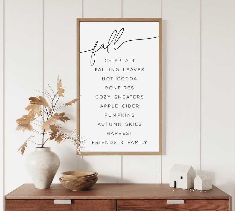 May include: A framed print with the word "fall" in cursive script and a list of fall-related words, including crisp air, falling leaves, hot cocoa, bonfires, cozy sweaters, apple cider, pumpkins, autumn skies, harvest, and friends & family.