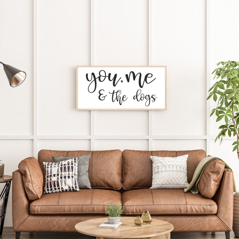 May include: A rectangular wall sign with a light wood frame featuring the black text "you, me & the dogs" in a cursive script on a white background. The sign is mounted on a white paneled wall above a brown leather sofa with decorative pillows.