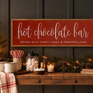 May include: A rectangular wooden sign with a red background and a light wood frame. The text reads "hot chocolate bar" in white script and "SERVED WITH CANDY CANES & MARSHMALLOWS" in white block letters. The sign is mounted on a dark wall.