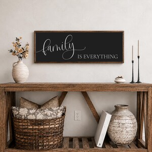 May include: A rectangular wall sign with a dark charcoal background and a wooden frame. The sign features the white cursive text "family" followed by the white block-letter text "IS EVERYTHING" centered horizontally on the surface of the sign.