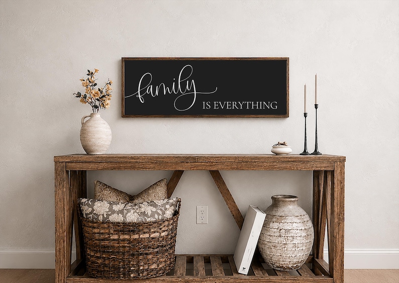 May include: A rectangular wall sign with a dark charcoal background and a wooden frame. The sign features the white cursive text "family" followed by the white block-letter text "IS EVERYTHING" centered horizontally on the surface of the sign.