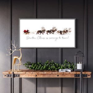 Puede incluir: Un cuadro rectangular con marco que presenta un fondo blanco con una ilustraci&oacute;n de Santa Claus en un trineo tirado por renos. El texto "Santa Claus is coming to town!" est&aacute; impreso en letra cursiva negra debajo del trineo.