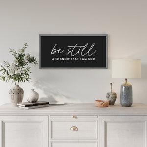 May include: A rectangular wall sign with a dark gray frame and a black background. The text "be still" is written in a white cursive script above the text "AND KNOW THAT I AM GOD" in a white sans-serif font, centered on the sign.