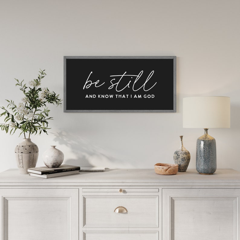 May include: A rectangular wall sign with a dark gray frame and a black background. The text "be still" is written in a white cursive script above the text "AND KNOW THAT I AM GOD" in a white sans-serif font, centered on the sign.