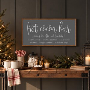 May include: A rectangular wall sign with a dark gray background and a light wood frame. The text reads: hot cocoa bar, warm up here, made fresh daily, marshmallows, crushed cookies, candy canes, sprinkles, whipped cream, chocolate chips.