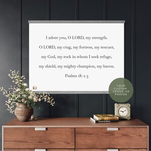 May include: A white rectangular wall hanging with a grey wooden frame at the top and bottom. The text reads: I adore you, O LORD, my strength. O LORD, my crag, my fortress, my rescuer, my God, my rock in whom I seek refuge, my shield, my mighty champion, my haven. Psalms 18: 2-3.