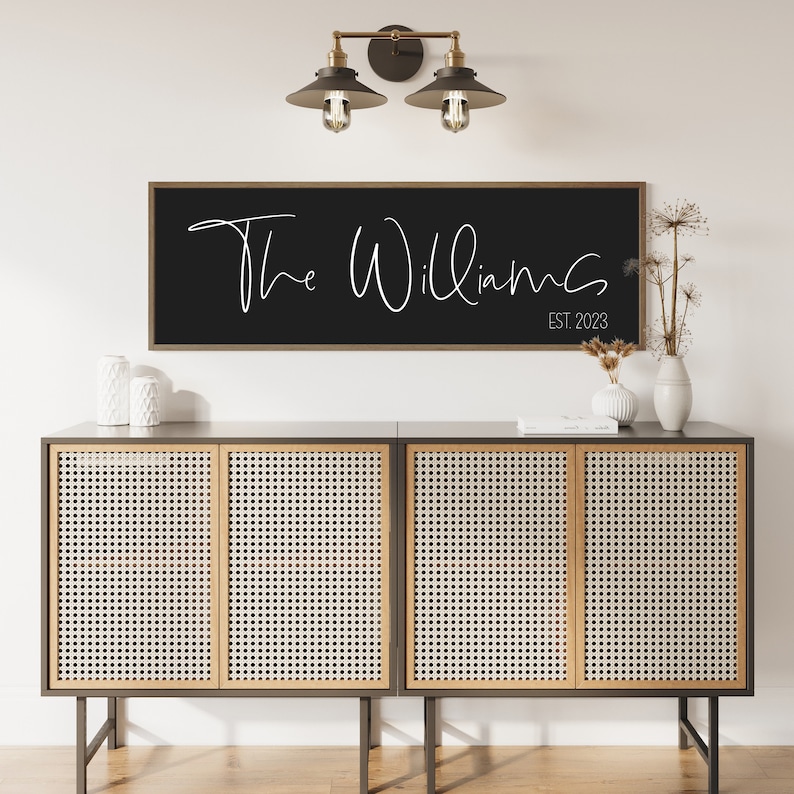 May include: A rectangular wall sign with a dark frame and a black background featuring the white text The Williams and EST. 2023 in a script and sans-serif font. The sign is mounted horizontally on a wall above a four-door cabinet with cane panels.