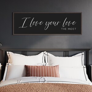 May include: A black framed sign with white calligraphy that reads "I love your love the most" hanging above a bed with white bedding and a striped throw pillow.