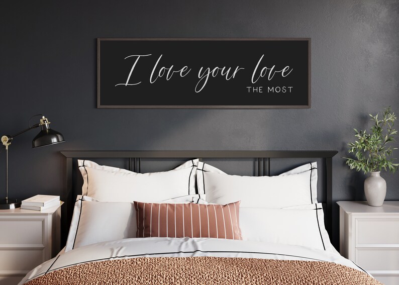 May include: A black framed sign with white calligraphy that reads "I love your love the most" hanging above a bed with white bedding and a striped throw pillow.
