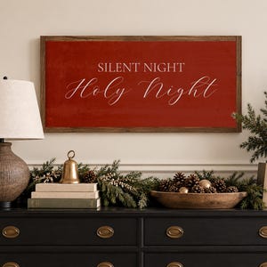 May include: A rectangular wall sign with a dark wood frame and a deep red background. The text "SILENT NIGHT" is printed in a white sans-serif font, and the words "Holy Night" are written below in a white, elegant, flowing script font.