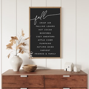 May include: A black framed print with white text that reads "fall" and lists the following: crisp air, falling leaves, hot cocoa, bonfires, cozy sweaters, apple cider, pumpkins, autumn skies, harvest, friends & family.