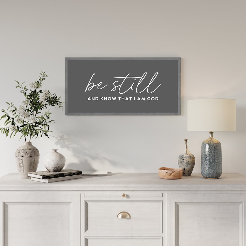 May include: A rectangular wall sign with a dark gray background and a light gray wooden frame. The sign features the white text "be still" in a cursive script font above the text "AND KNOW THAT I AM GOD" in a clean, sans-serif capital font.
