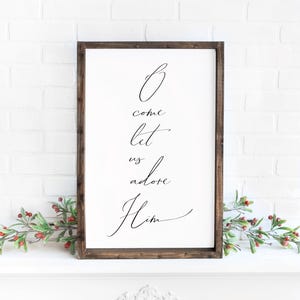 May include: A framed white sign with the words "O come let us adore Him" in elegant black script. The wooden frame is dark brown. Decorative greenery with red berries accents the bottom.