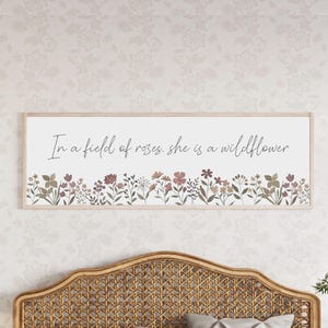 Puede incluir: Un cuadro rectangular con marco de madera clara que presenta el texto "In a field of roses, she is a wildflower" en letra cursiva negra sobre una ilustraci&oacute;n horizontal de flores silvestres en tonos apagados de rosa, marr&oacute;n y verde.