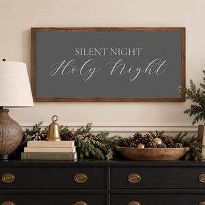 May include: A rectangular wall sign with a dark wood frame and a dark gray background featuring the white text SILENT NIGHT Holy Night in a combination of block and script fonts, centered horizontally on the wall above a black wooden dresser.