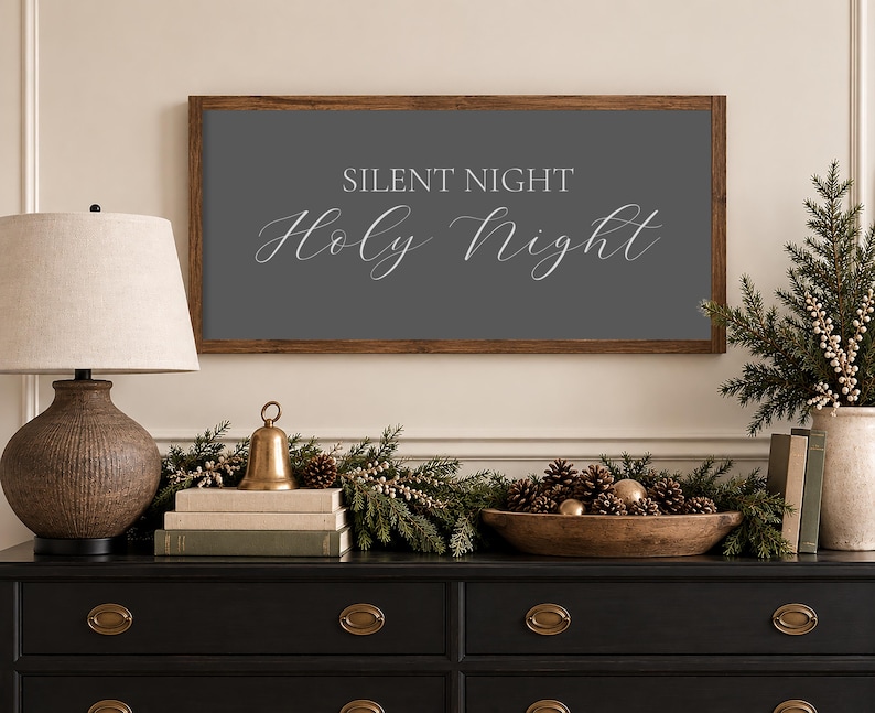 May include: A rectangular wall sign with a dark wood frame and a dark gray background featuring the white text SILENT NIGHT Holy Night in a combination of block and script fonts, centered horizontally on the wall above a black wooden dresser.