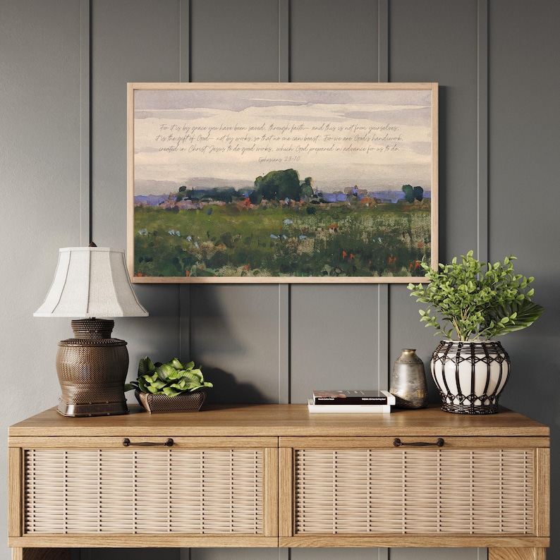 May include: A framed landscape print with a light wood frame hangs above a wooden console table. The print depicts a field with trees and a quote from Ephesians 2:8-10. A lamp, plants, and decorative items are on the table.
