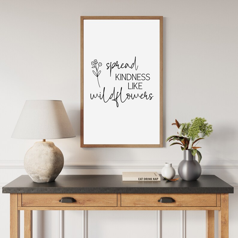 以下が含まれることがあります： 「Spread Kindness Like Wildflowers」の文字が黒字で書かれた額入りのプリント。茶色の木製フレームに入っており、ランプと花瓶のある木製のテーブルに飾られています。