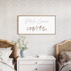 May include: A rectangular wall sign with a light wood frame featuring the name Mila Grace in black cursive script above a row of small, muted-coloured botanical illustrations including various flowers and leaves on a white background.