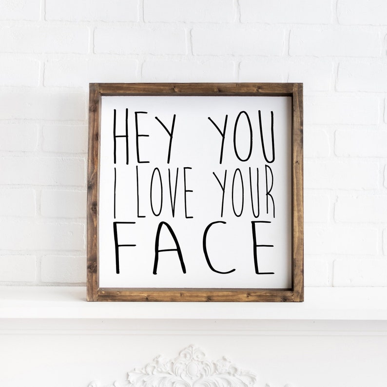 K&ouml;nnte beinhalten: Ein wei&szlig;es Holzschild mit einem braunen Holzrahmen. Das Schild sagt "Hey You I Love Your Face" in schwarzer handschriftlicher Schrift.
