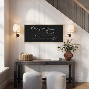 May include: A rectangular black wall sign with a light wood frame featuring white cursive and print text that reads: Our family is a circle of strength and love, founded on faith, joined in love, kept by God. The sign is mounted on a white wall.