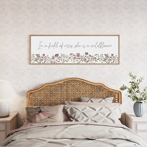 Puede incluir: Un letrero de pared rectangular con un marco de madera clara que presenta el texto cursivo negro In a field of roses, she is a wildflower sobre una colorida ilustraci&oacute;n de varias flores silvestres peque&ntilde;as en tonos rosa, verde y marr&oacute;n.