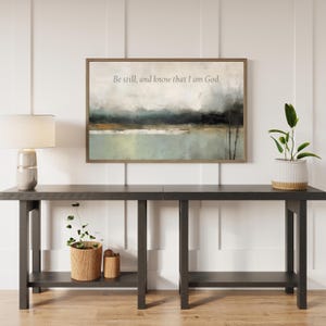 May include: An abstract landscape painting in a wooden frame with the text "Be still, and know that I am God." The painting hangs above a dark gray console table with a lamp and plants.