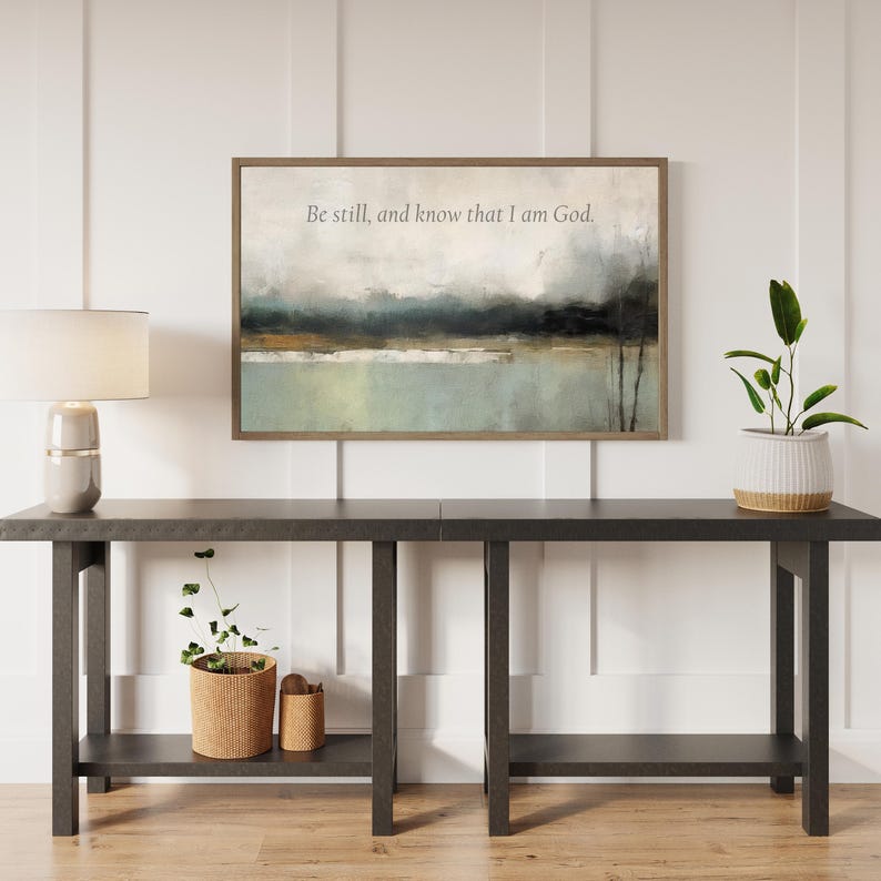 May include: An abstract landscape painting in a wooden frame with the text "Be still, and know that I am God." The painting hangs above a dark gray console table with a lamp and plants.