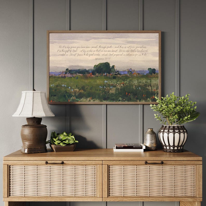 May include: A framed landscape painting with a field of green and wildflowers, and a quote in script. The painting hangs above a wooden cabinet with a lamp, a plant, and decorative items. The cabinet has a woven design.