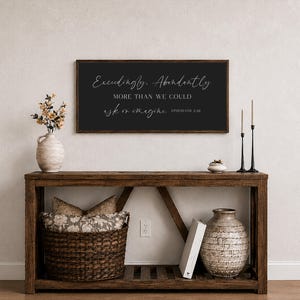 Op de afbeelding: Een houten console met een donker omlijst bord met de tekst "Exceedingly, Abundantly, More than we could ask or imagine, Ephesians 3:20." De tafel heeft een gevlochten mand, een vaas en decoratieve items.