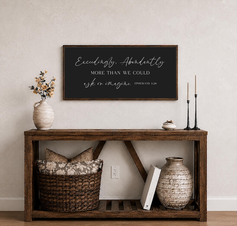 Op de afbeelding: Een houten console met een donker omlijst bord met de tekst "Exceedingly, Abundantly, More than we could ask or imagine, Ephesians 3:20." De tafel heeft een gevlochten mand, een vaas en decoratieve items.