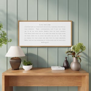 May include: A framed rectangular sign with the text "Love the Lord" in a white font. The sign is displayed on a wooden table with a lamp, plants, and decorative items. The background is a light green wall.