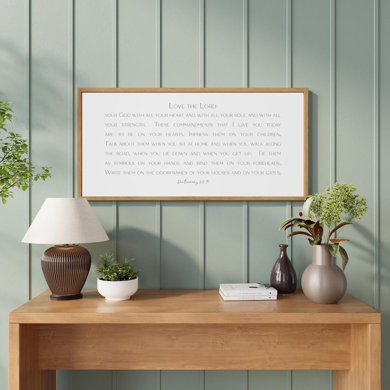 May include: A framed rectangular sign with the text "Love the Lord" in a white font. The sign is displayed on a wooden table with a lamp, plants, and decorative items. The background is a light green wall.