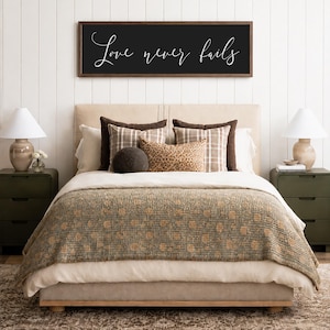 May include: A bedroom scene featuring a bed with a beige headboard, layered bedding, and decorative pillows. A dark wooden framed sign above the bed reads "Love never fails" in white script. Two green nightstands with lamps flank the bed.