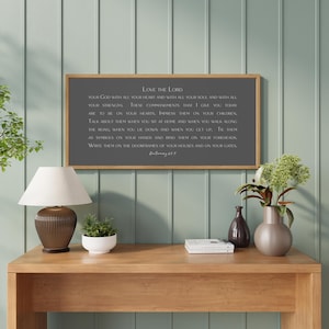 May include: A framed wall art piece with the text "Love the Lord" in white on a gray background. The wooden frame surrounds the text, which is a religious quote. The artwork is displayed above a wooden table.