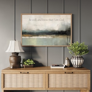 May include: A framed landscape art print with the text "Be still, and know that I am God." The artwork is displayed above a wooden console table with a lamp, potted plants, and decorative items. The color palette includes muted greens, blues, and browns.