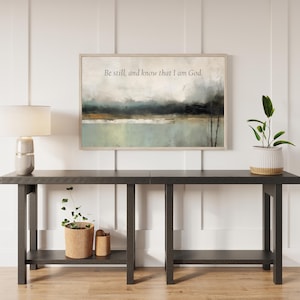 May include: A framed landscape art print with the text "Be still, and know that I am God." The artwork features a muted color palette of blues, greens, and browns. The print is displayed above a dark gray console table with a lamp and plants.