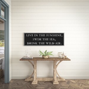May include: A black framed sign with white text that reads "Live in the sunshine, swim the sea, drink the wild air." The sign is hanging on a white shiplap wall above a wooden console table.