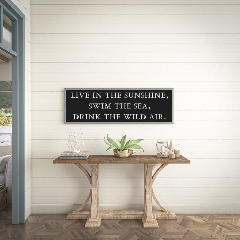 May include: A black framed sign with white text that reads "Live in the sunshine, swim the sea, drink the wild air." The sign is hanging on a white shiplap wall above a wooden console table.