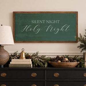 May include: A rectangular wall sign with a dark green background and a brown wooden frame. The text SILENT NIGHT is printed in a simple sans-serif font, while the words Holy Night are written below in a white, elegant, flowing script font.