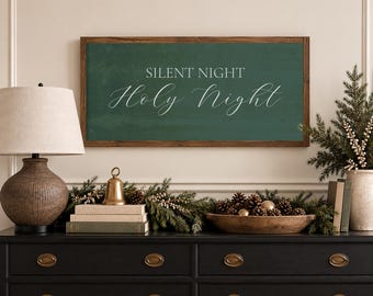 May include: A rectangular wall sign with a dark green background and a brown wooden frame. The text SILENT NIGHT is printed in a simple sans-serif font, while the words Holy Night are written below in a white, elegant, flowing script font.