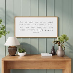 May include: A rectangular wall sign with a light wood frame and a white background featuring black text that reads: And we know that in all things God works for the good of those who love him, who have been called according to His purpose. Romans 8:28.